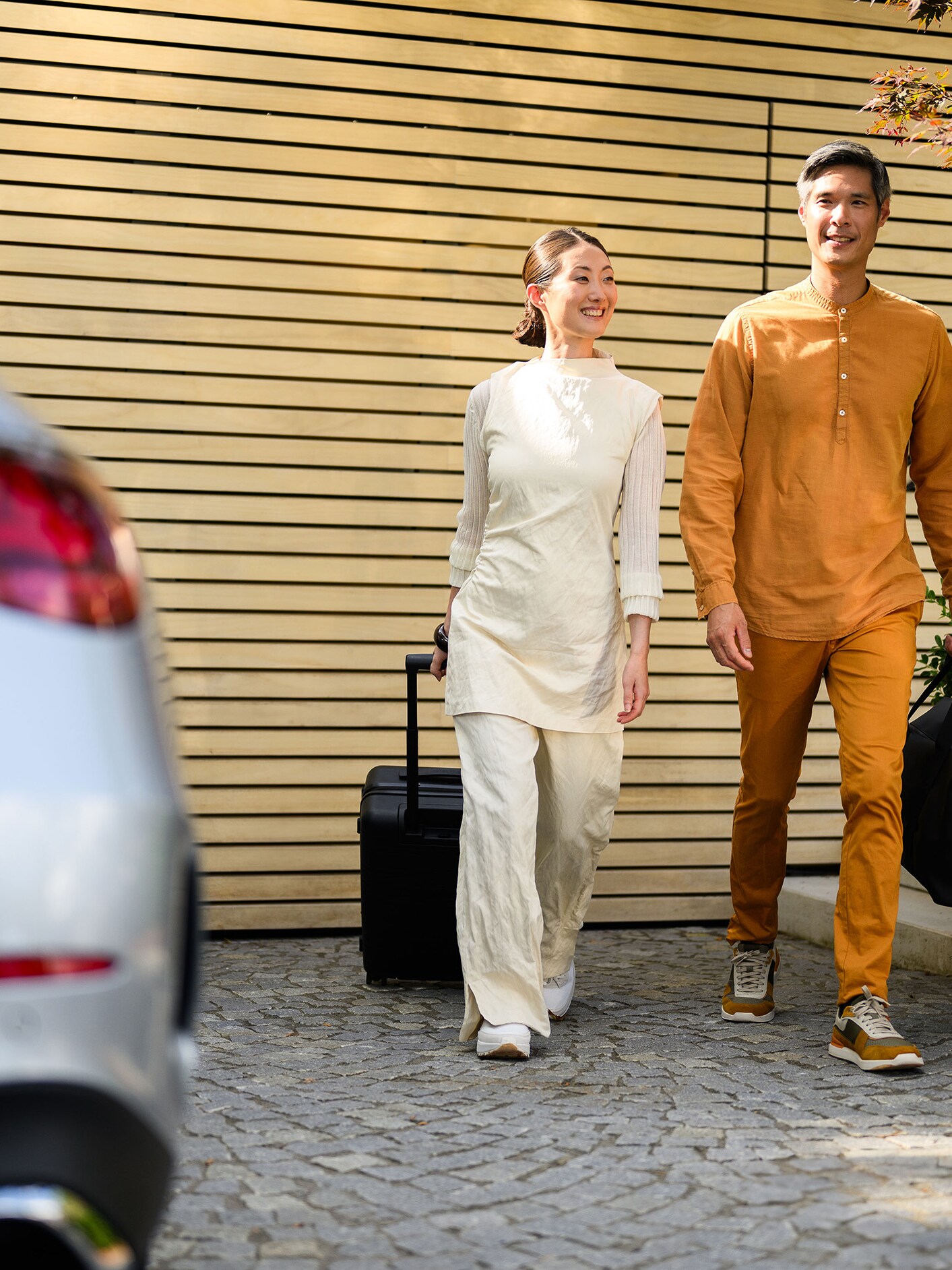 Two people walking to a Mercedes-Benz vehicle, one walking with a suitcase. 兩個人走向賓士車，一位拉著行李箱