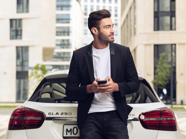 一位男仕靠在他的 Mercedes-Benz GLA 並透過他智慧型手機上的 Mercedes me 應用程式和導航主題數位增訂查詢抵達地點的天氣。